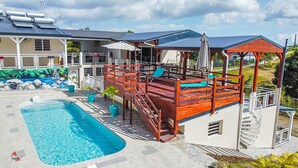 A swimming pool with a wooden deck and lounge chairs, a covered seating area, and a building with a mural. - Villa Angerville, spacious villa that can accommodate up to 24 people (Le Moule)
