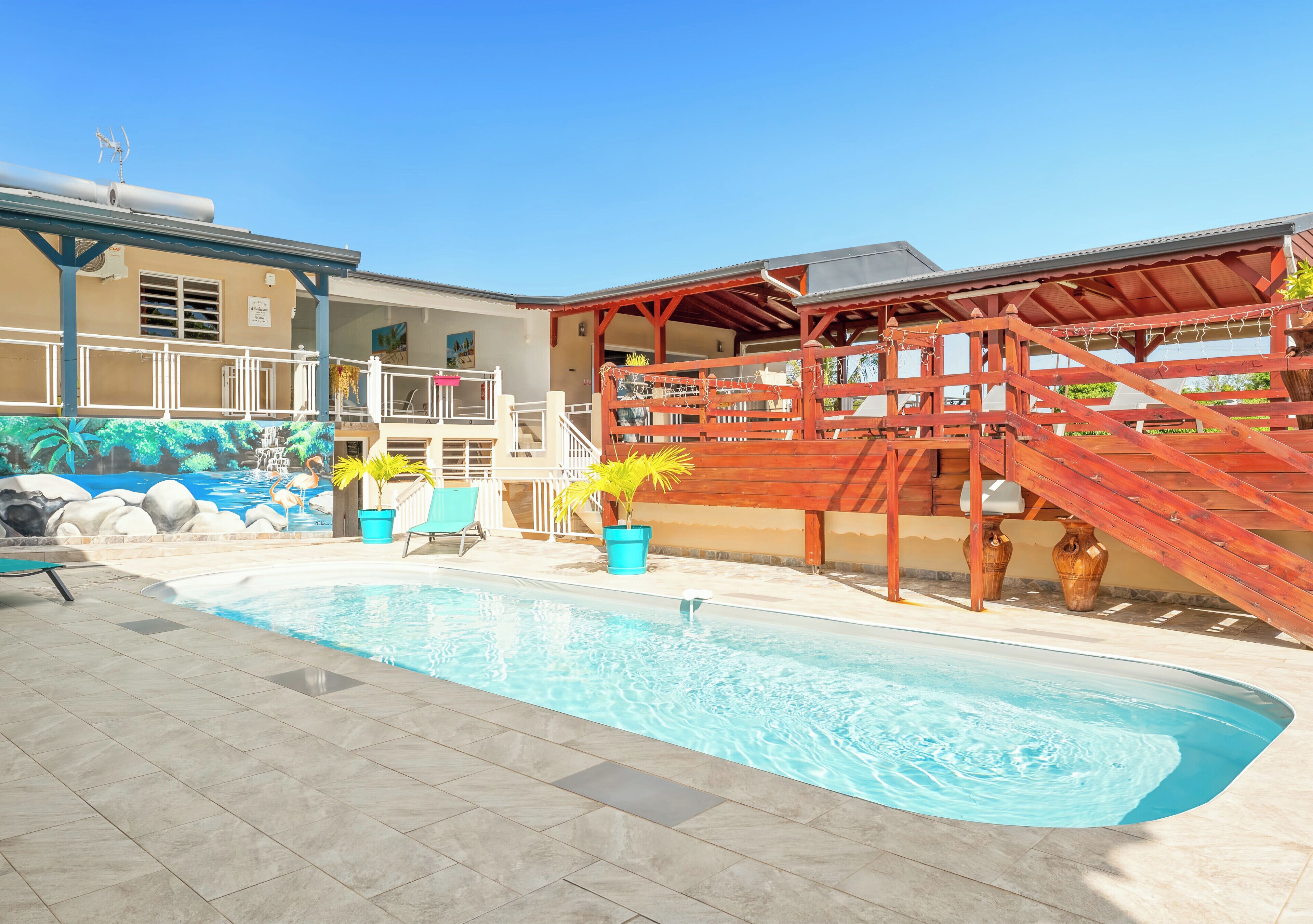 Una piscina con muelle de madera, sillas reclinables, un mural y una zona cubierta con escaleras.