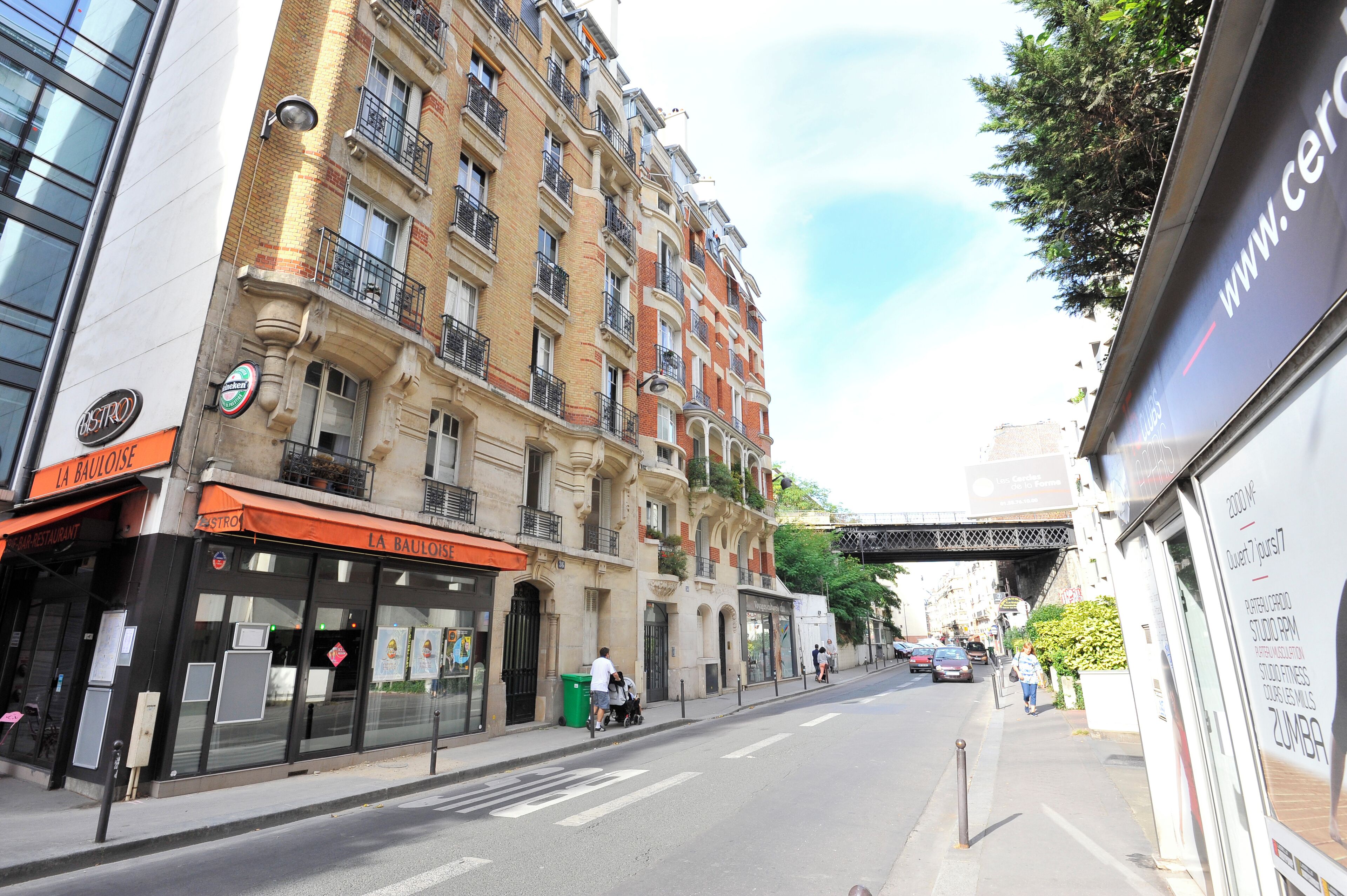 Straßenansicht mit einem Restaurant namens "La Bauloise" auf der linken Seite, einem mehrstöckigen Gebäude mit Balkonen und einem Fußweg.