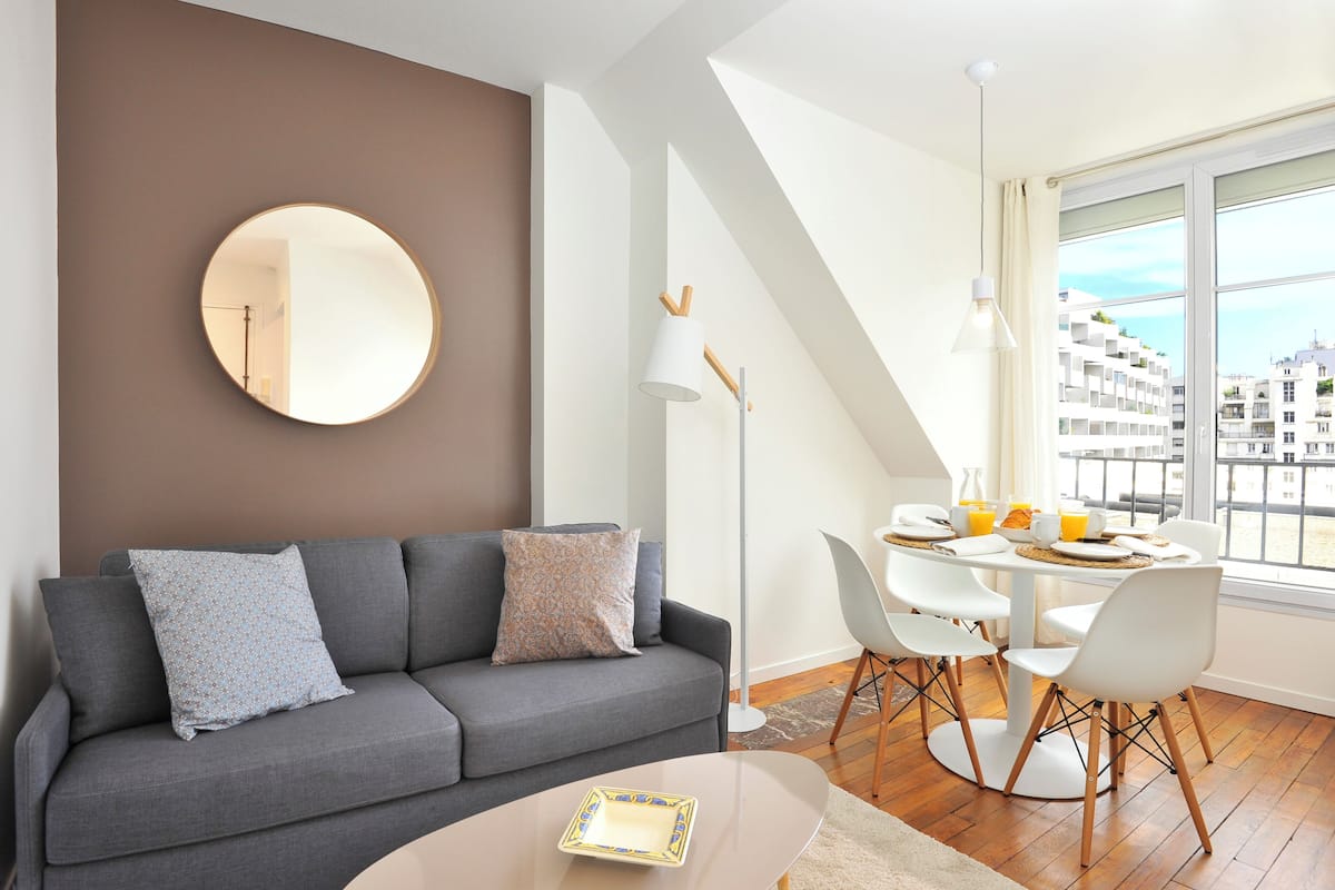 A modern living room with a grey sofa, a round mirror, and a dining area with a table set for breakfast.