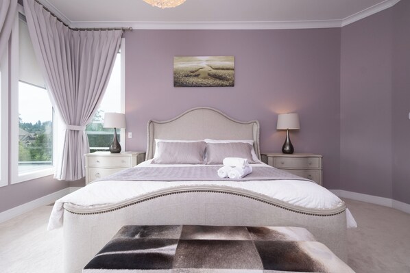 A bedroom with a large bed, two bedside tables, a nightstand, a window with curtains, and a painting on the wall.