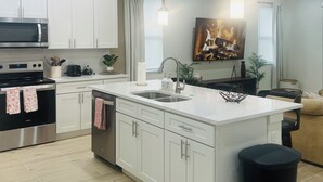 A modern kitchen with white cabinetry, a stainless steel oven, a microwave, and a large island with a sink. There are pendant lights above the island and a painting on the wall. - Meet. Play. Stay. • Relax & Play in Orlando (Orlando)