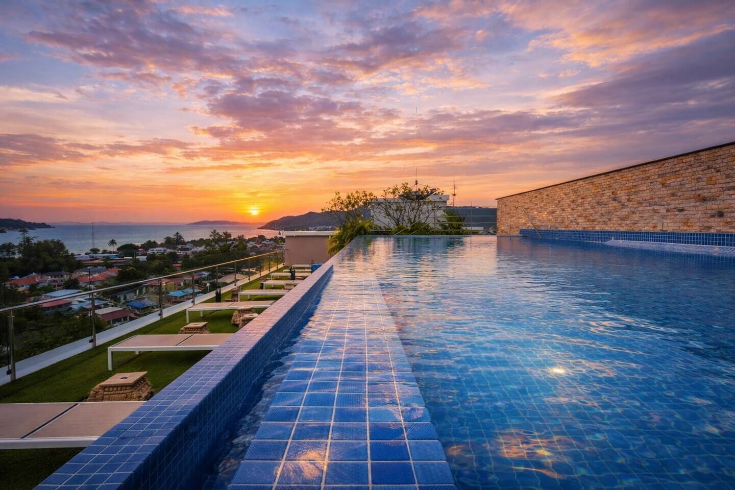 A swimming pool with a view of the sunset over the ocean, a stone wall, and a grassy area with a small building.