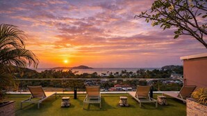 Eine Terrasse mit Liegestühlen, Blick auf den Sonnenuntergang über dem Meer und eine ferne Stadtansicht.