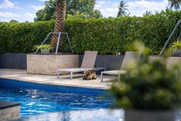 A swimming pool with lounge chairs and a small seating area, surrounded by a hedge and a palm tree.