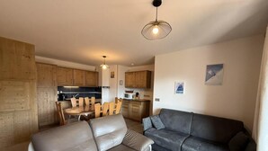 A modern living room with a grey sofa, a dark grey couch, and a wooden coffee table. There is a dining area with a wooden table and chairs, and a kitchen in the background. - 250m from ski slopes, 1st floor, view resort, terrace, parking, tv, ski locker, 53m², Les Saisies (Villard-sur-Doron)