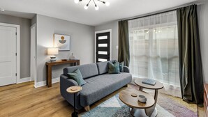 Ein modernes Wohnzimmer mit einem grauen Sofa, einem kleinen runden Tisch, einer Lampe und einem großen Fenster mit Vorhängen.