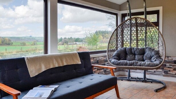Un salon moderne avec un canapé et un fauteuil suspendu, de grandes fenêtres donnant sur un paysage verdoyant et un mur en pierre.