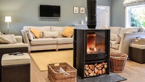 Un salon chaleureux avec un poêle à bois noir, un canapé gris, des paniers en osier et un téléviseur à écran plat fixé au mur.