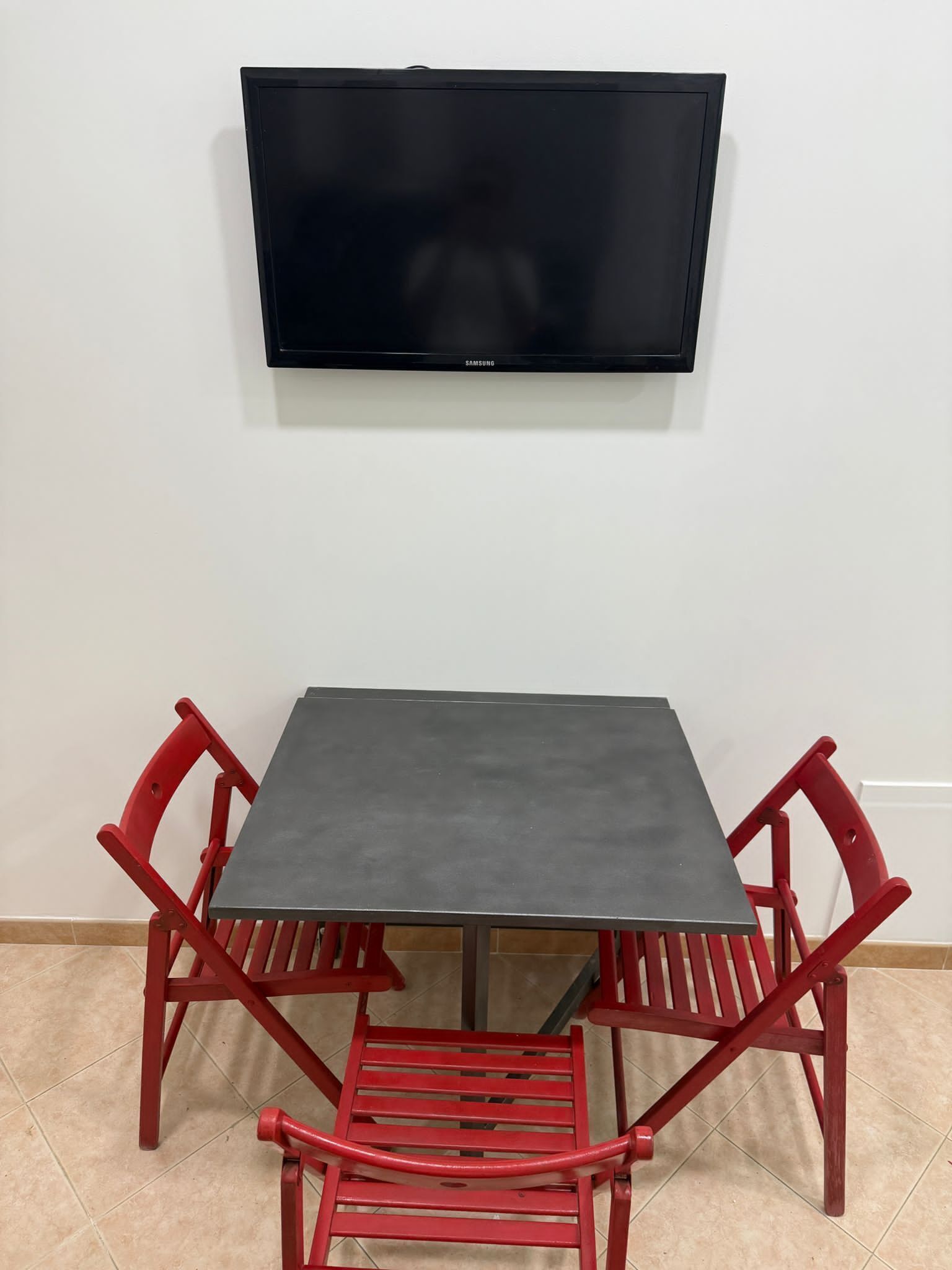Een kamer met een flatscreen televisie aan de muur, een kleine tafel en twee rode stoelen.