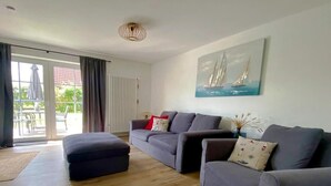 A living room with a grey sofa set, a wooden floor, a large window with curtains, and a sailboat painting on the wall. - Ostfreesenstuuv max. 4 person in the heart of Neßmersiel Terrace, garden, WLAN- Wallbox (Nessmersiel)