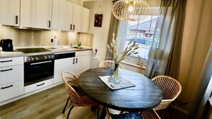A modern kitchen with white cabinets, a black countertop, and a dining area with wicker chairs and a round table. - Ostfreesenstuuv max. 4 person in the heart of Neßmersiel Terrace, garden, WLAN- Wallbox (Nessmersiel)