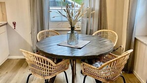 A dining area with a round table, wicker chairs, a vase with dried plants, and a hanging light fixture. - Ostfreesenstuuv max. 4 person in the heart of Neßmersiel Terrace, garden, WLAN- Wallbox (Nessmersiel)