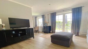 A spacious living room with a flat-screen TV on a black cabinet, a grey ottoman, and a dining area with a table and chairs. - Ostfreesenstuuv max. 4 person in the heart of Neßmersiel Terrace, garden, WLAN- Wallbox (Nessmersiel)