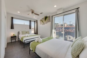 Una habitación de hotel moderna con dos camas, una mesita de noche con lámpara, ventilador de techo y un ventanal grande con un cartel de 'Welcome'.