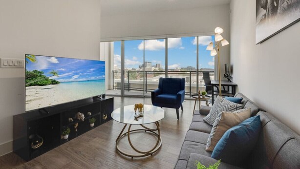 Uma sala de estar moderna com uma TV grande, uma poltrona azul, uma mesa de centro de vidro e um sofá cinza.