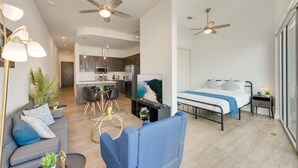 Um apartamento moderno com sala de estar, área de jantar, cozinha e quarto. Possui ventiladores de teto, TV de tela plana, cama com cobertor azul e uma pequena mesa de cabeceira com abajur.