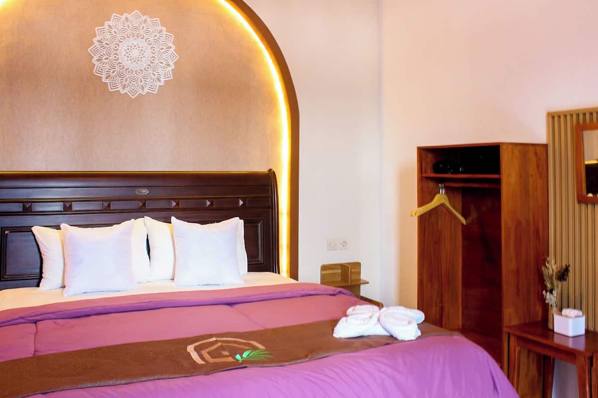 Una cama bien hecha con almohadas blancas y una colcha púrpura, un cabecero de madera con un diseño decorativo, y un armario de madera con un espejo y una barra para colgar.