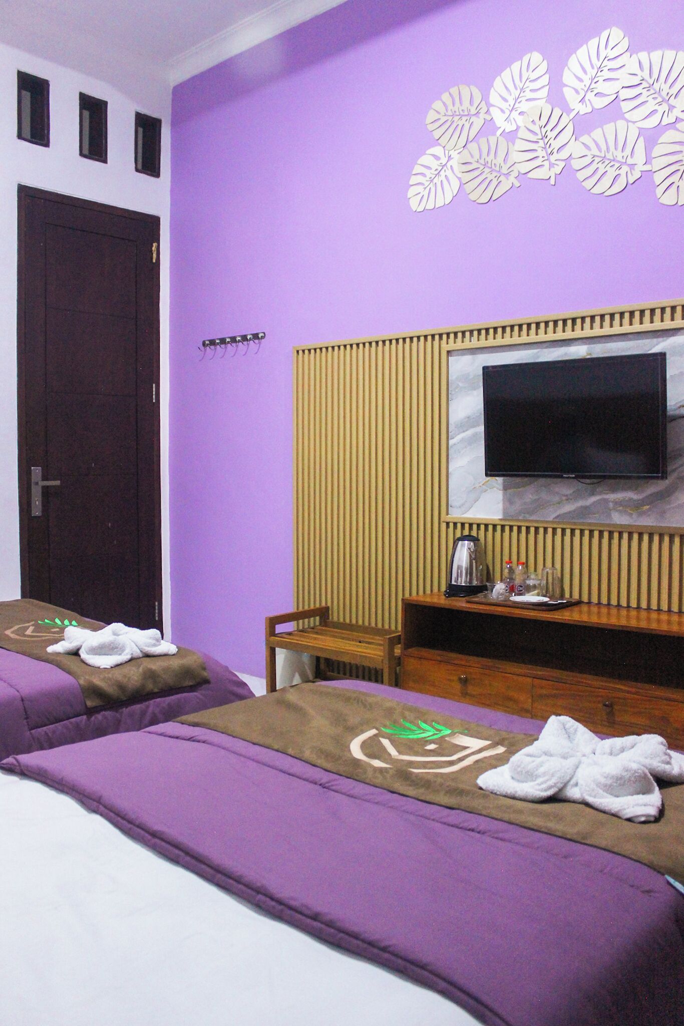 Habitación de hotel con dos camas, un televisor montado en un panel de madera, una pequeña mesa de madera con un hervidor y una pared con motivo de hojas.