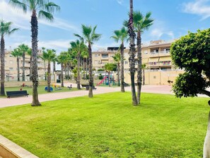 A park with palm trees, a playground, and a building with balconies in the background.