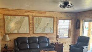 A cozy living room with wooden walls, two large maps, a leather sofa, a wooden coffee table, and a window with blinds. - Wake up to breathtaking sunrise views from the porch of this 2 BR waterview home (Sturgeon Bay)