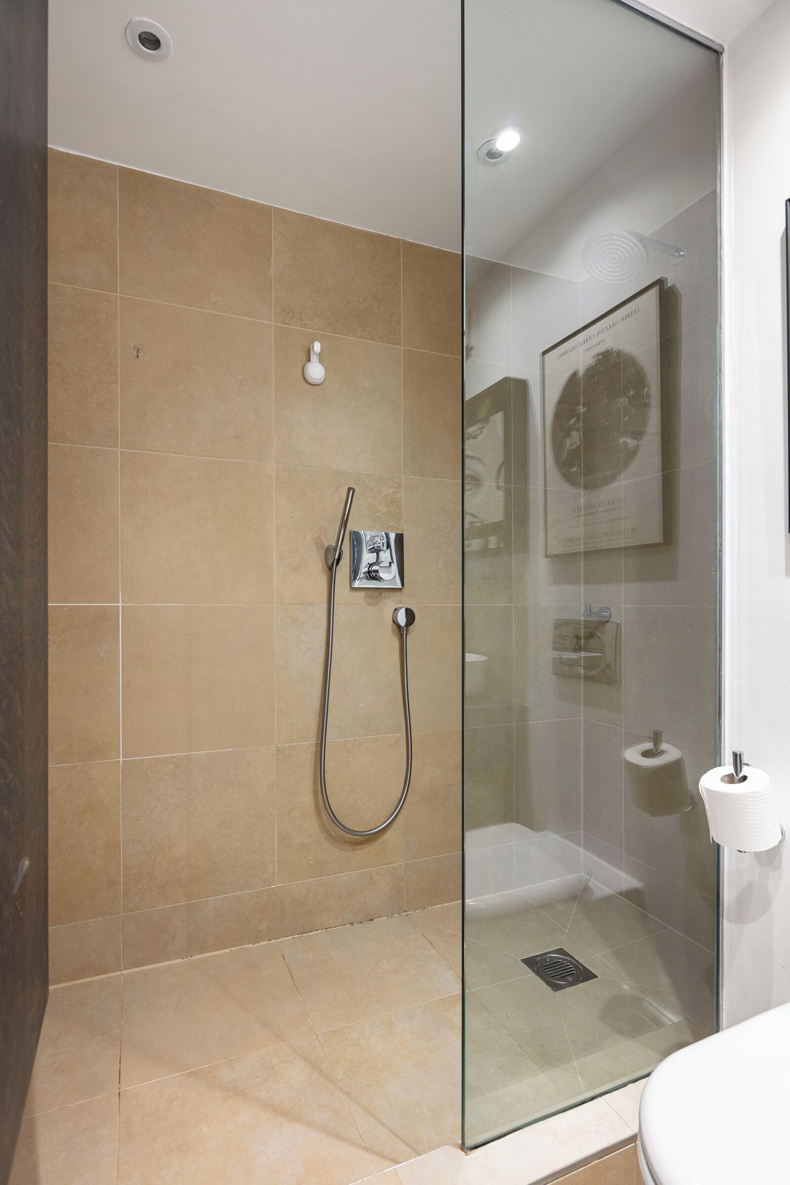 Een moderne badkamer met een douchecabine van glas, beige tegels, een toiletrolhouder en een lijst met een foto aan de muur.