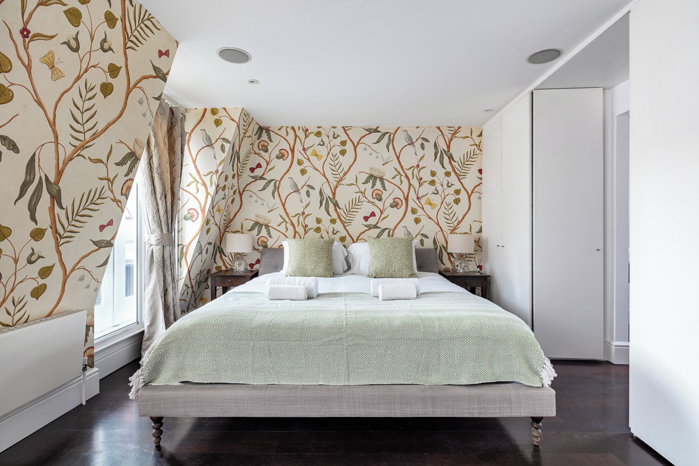 Un lit bien fait, recouvert d’une couvre-lit à motifs verts et blancs, avec deux oreillers de chaque côté, dans une pièce dont le papier peint est orné de motifs floraux et dont le plancher est en bois foncé.