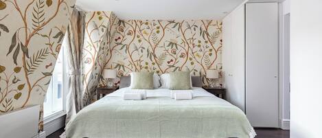 Un lit bien fait, recouvert d’une couvre-lit à motifs verts et blancs, avec deux oreillers de chaque côté, dans une pièce dont le papier peint est orné de motifs floraux et dont le plancher est en bois foncé.