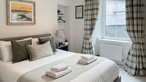 Een netjes opgemaakt bed met kussens en gevouwen handdoeken, een nachtkastje met een lamp en een klein plantje, en een raam met gordijnen.