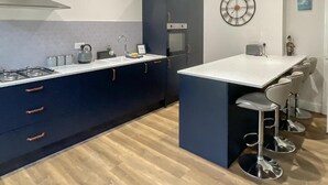 Een moderne keuken met donkerblauwe kastjes, een wit aanrechtblad en een ingebouwde oven. Er staan barkrukken en er hangt een wandklok.