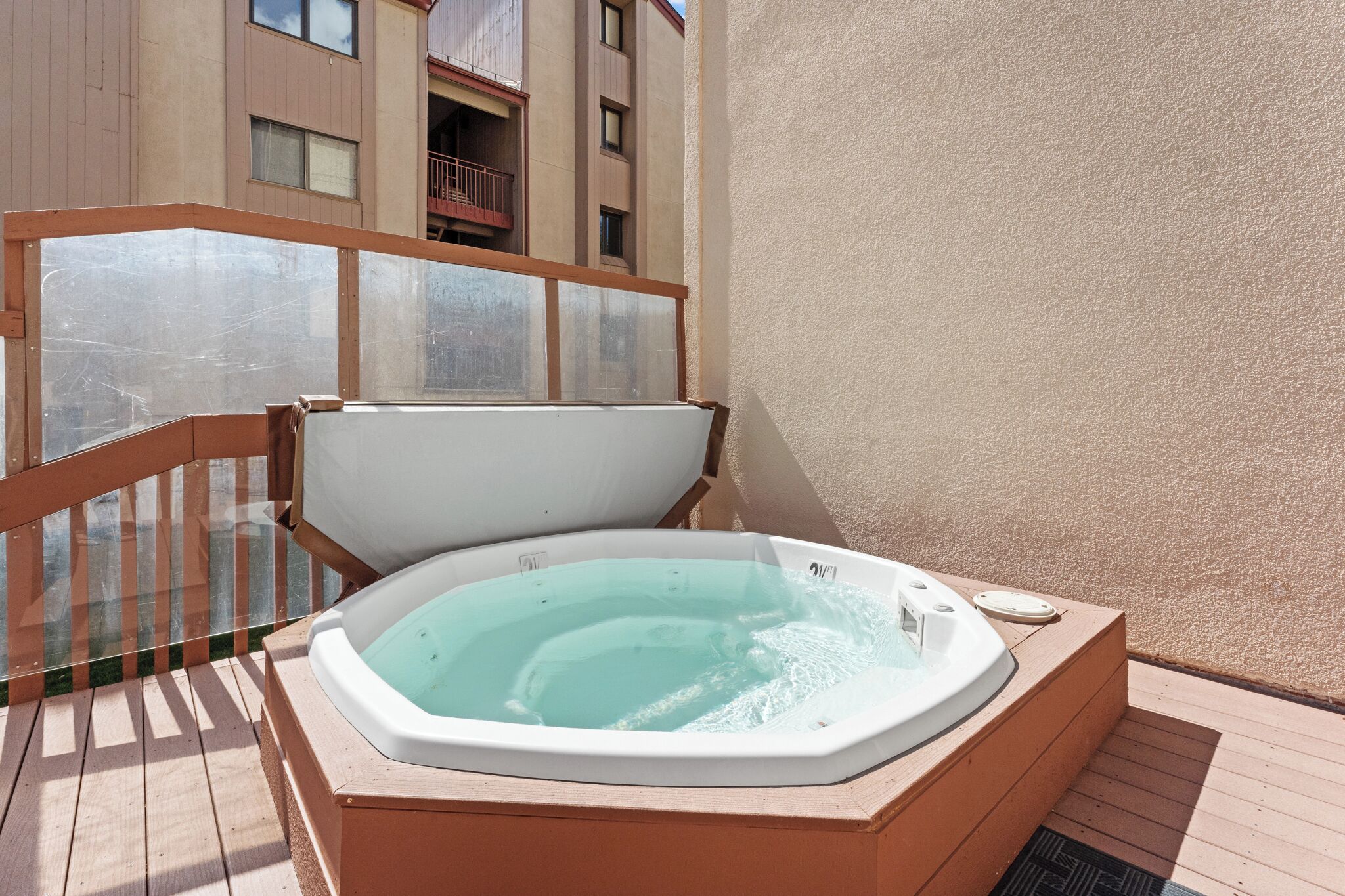 A hot tub on a wooden deck with a glass balustrade and a building in the background.