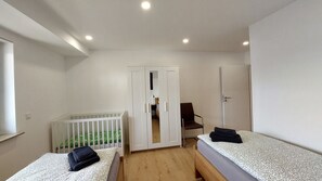 A small, modern room with two beds, a white crib, a chair, and a wardrobe. - New modern apartment "Gerda" for up to 6 people (Mörlenbach)