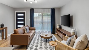 Una sala de estar moderna con un sofá de cuero marrón, una mesa de centro redonda, una silla de mimbre y un televisor de pantalla plana montado en la pared.