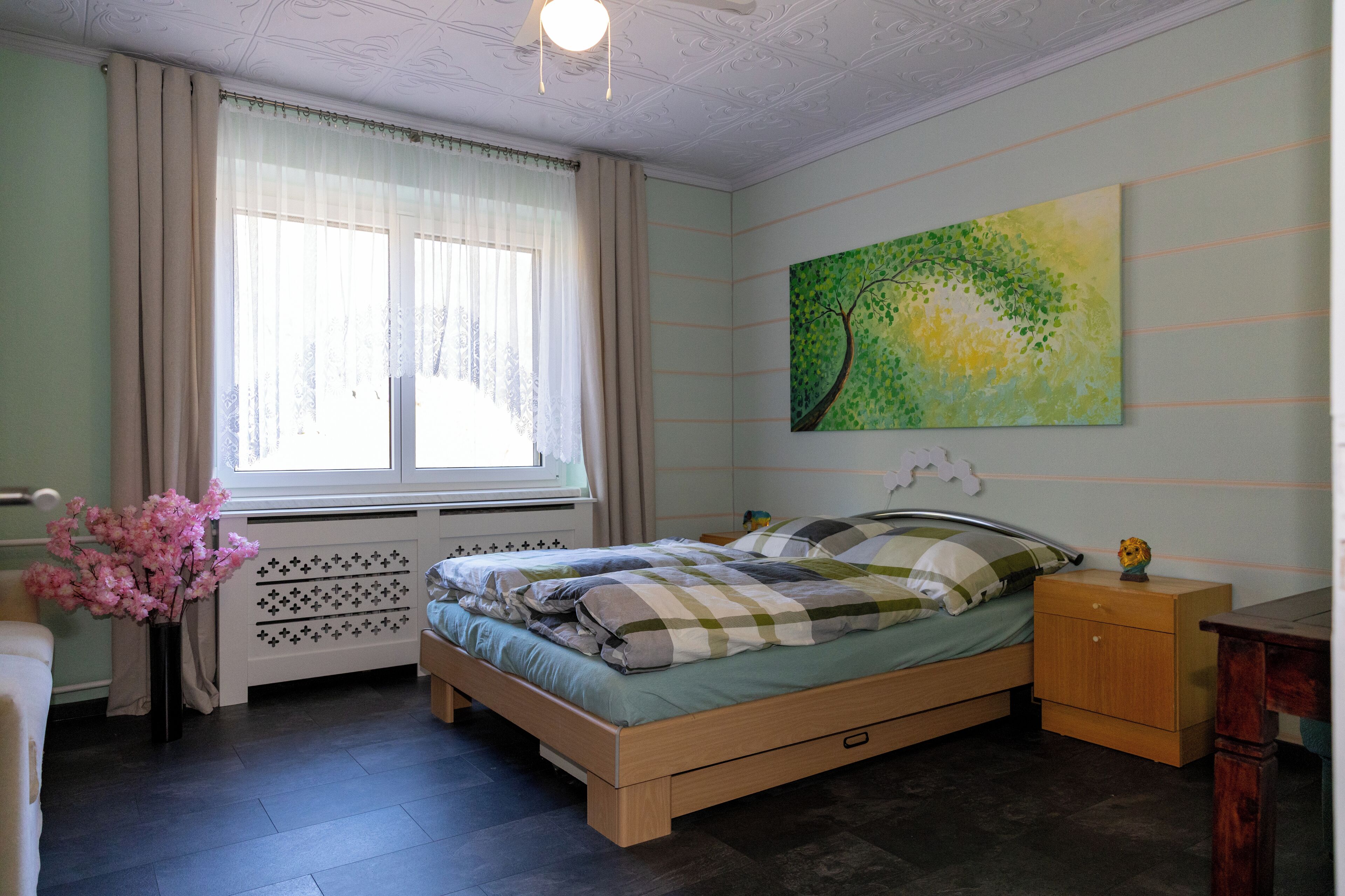 Ein Schlafzimmer mit Bett, Nachttisch, Kommode, Heizkörper, einem Fenster mit Vorhängen und einem Bild an der Wand.