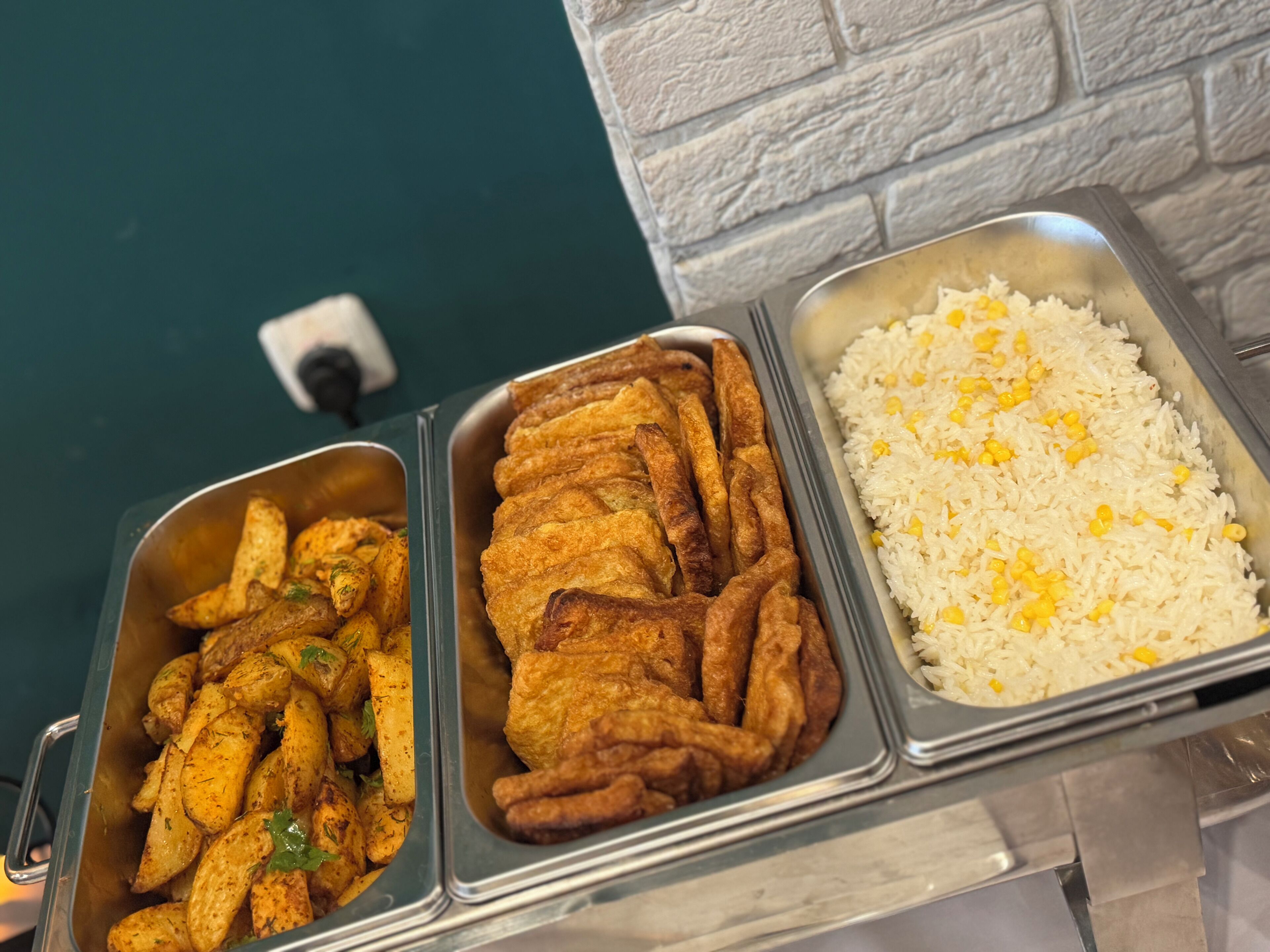 A buffet with three trays of food: fried chicken, seasoned potatoes, and white rice with corn.