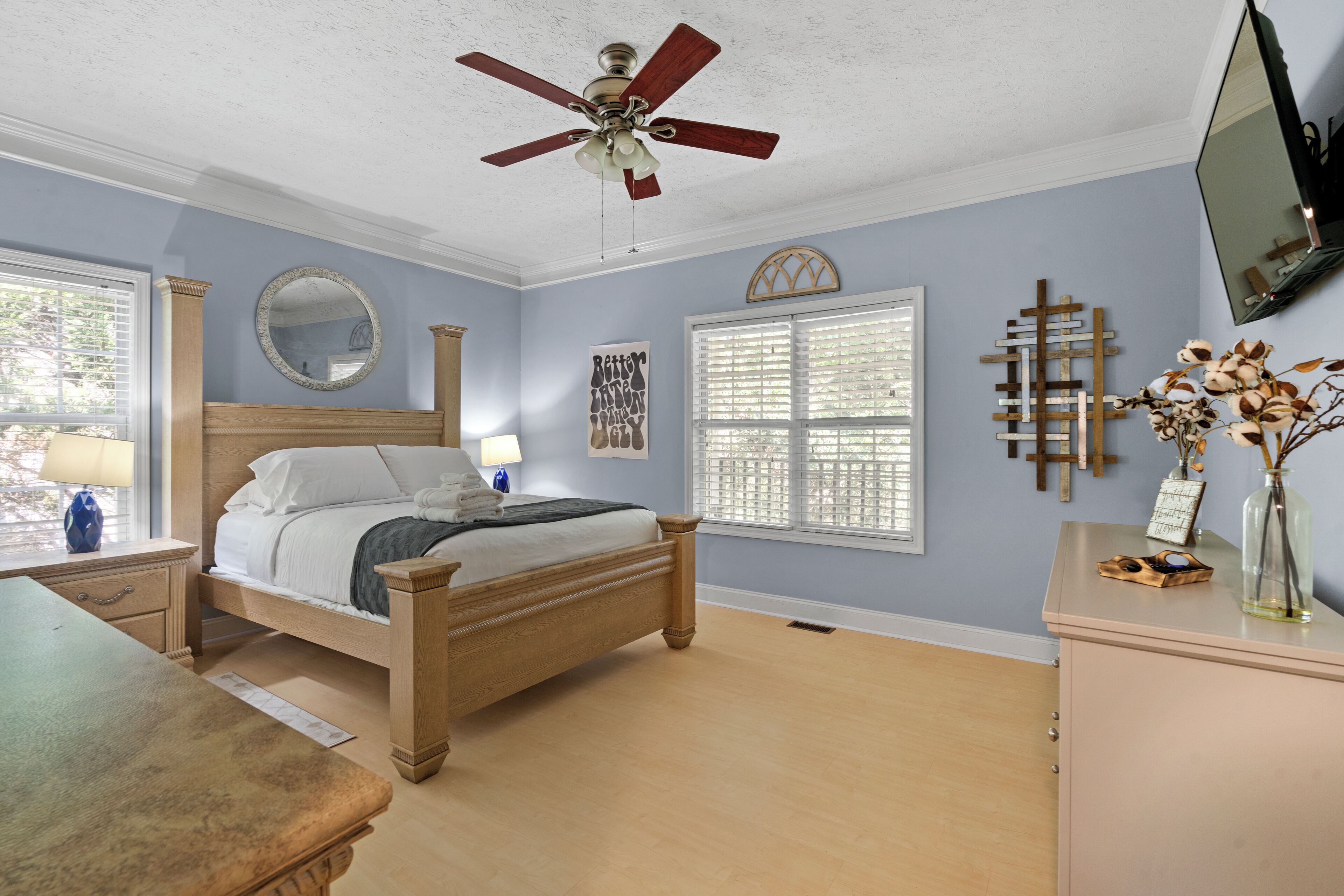 A bedroom with a bed, a desk, a dresser, a ceiling fan, and a large window.