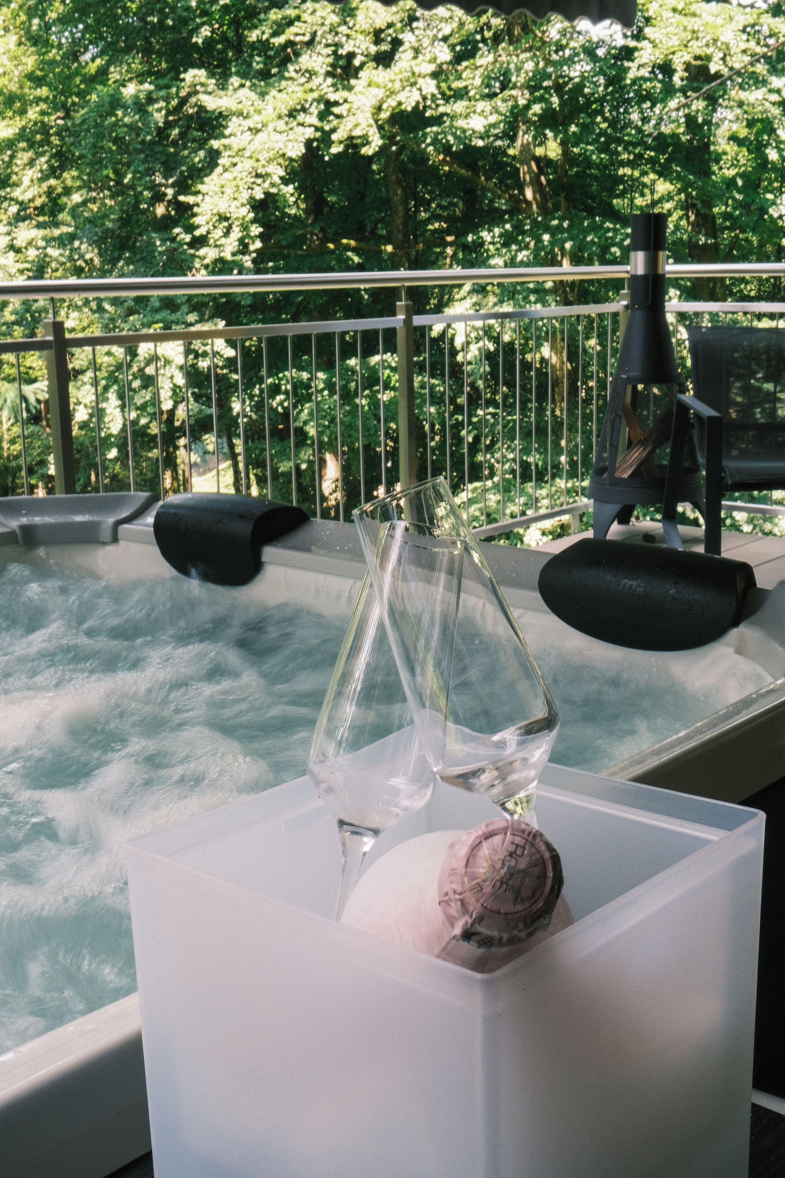 Ein Whirlpool mit einer klaren Glasvase und einem dekorativen Gegenstand auf einer weißen Plattform, umgeben von einem Metallgeländer und üppiger Vegetation im Hintergrund.