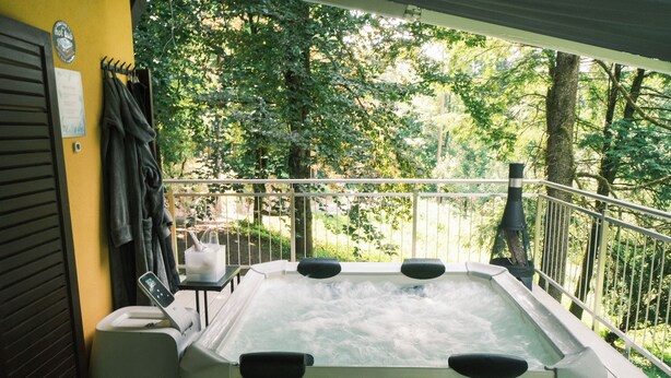 Un jacuzzi con cuatro chorros, un perchero y una mesita con una silla en un balcón con vistas a árboles.