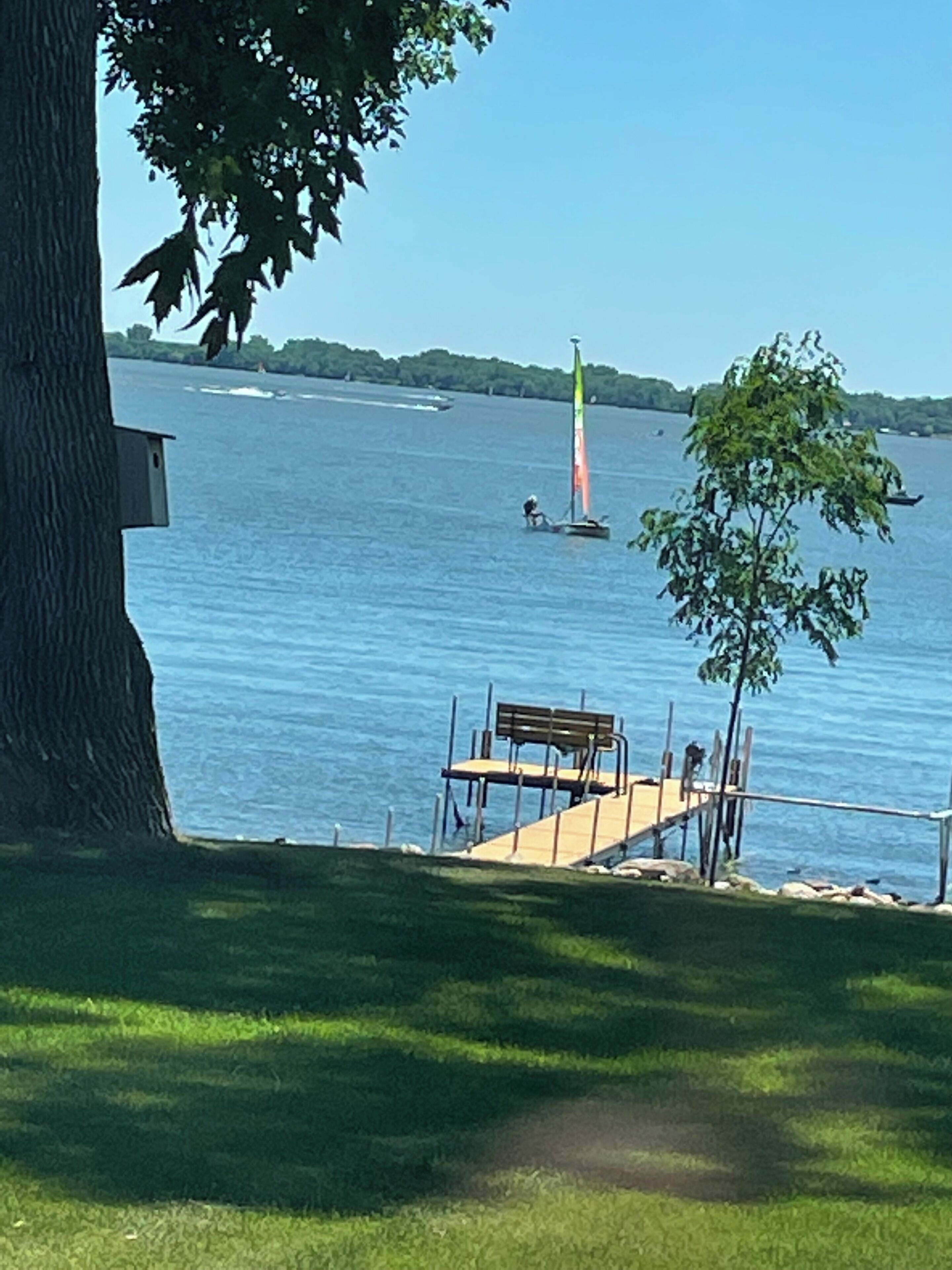 A lakeside view with a dock, a bench, and a sailboat in the distance.