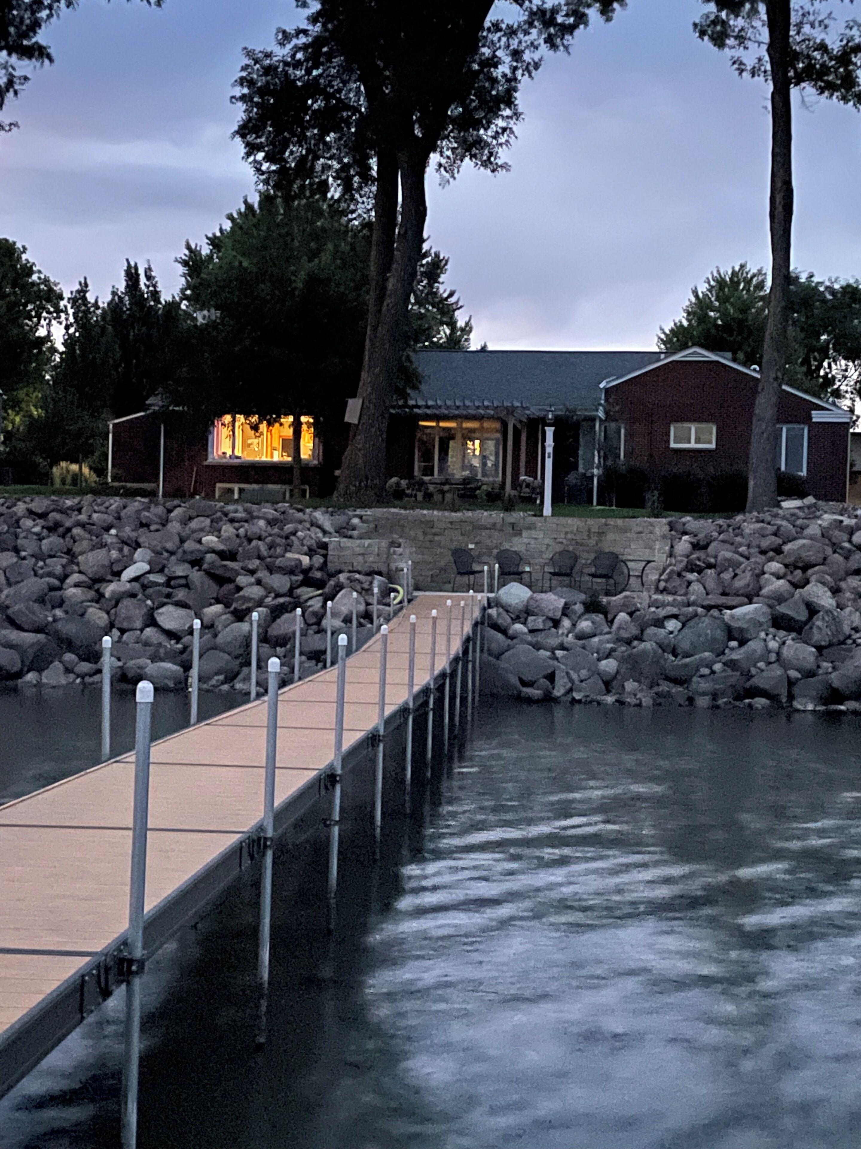 A wooden dock leading to a stone pier, with a house and trees in the background.