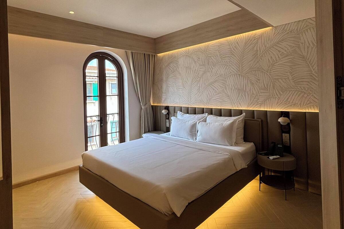 A modern hotel room with a large bed, a built-in headboard, and a view through a window with curtains.