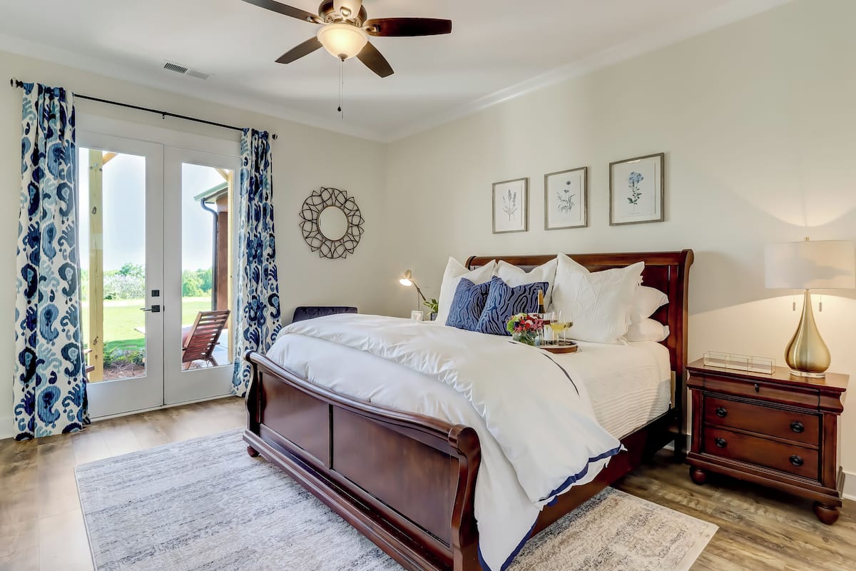 Quarto com cama grande, mesa de cabeceira de madeira, ventilador de teto e vista através de uma porta de vidro.