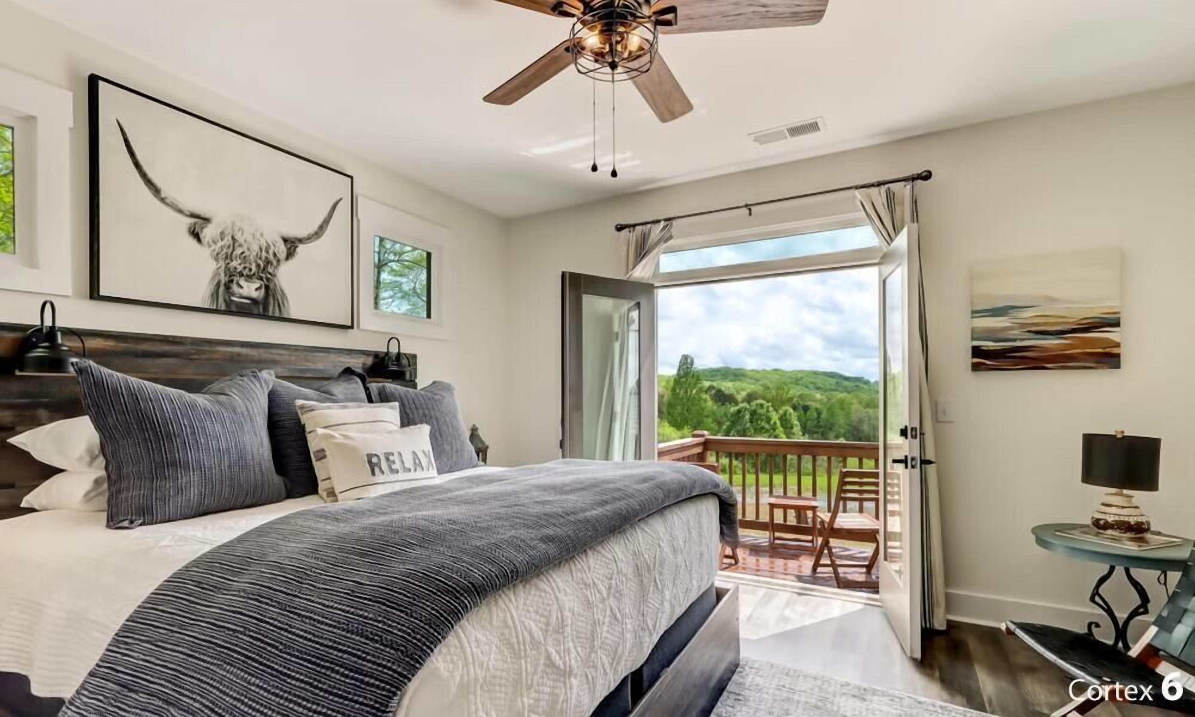 A bedroom with a large bed, a ceiling fan, a balcony with a view of greenery, and a painting on the wall.