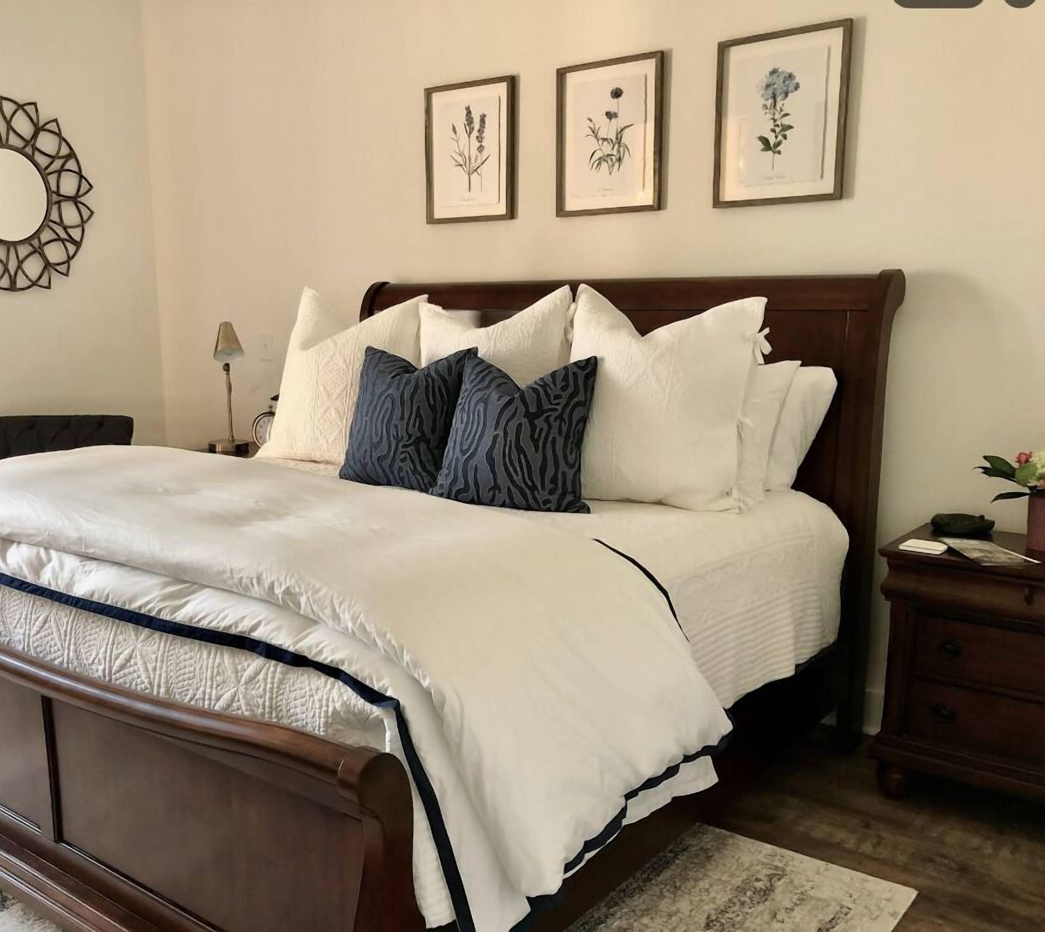 A neatly made bed with white and dark blue pillows, a wooden headboard, and framed botanical prints on the wall.