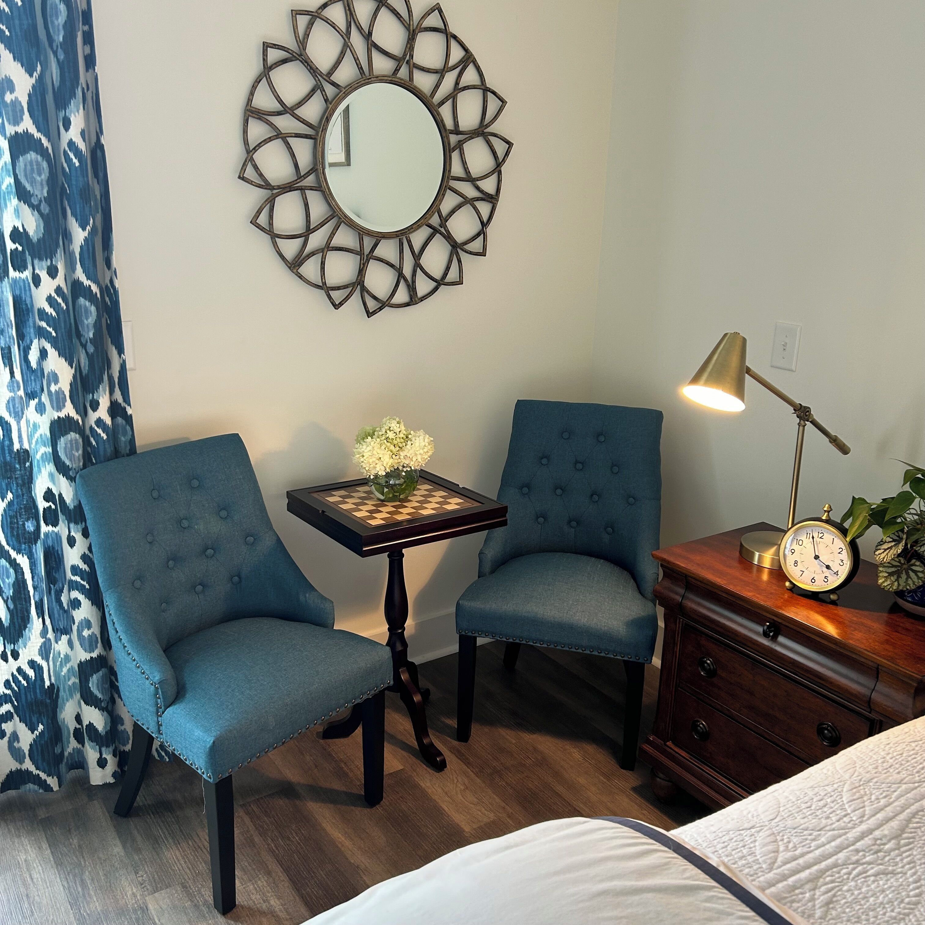 Una habitación con dos sillones azules, una mesita con un tablero de ajedrez, un espejo, una lámpara y un reloj sobre una cómoda.