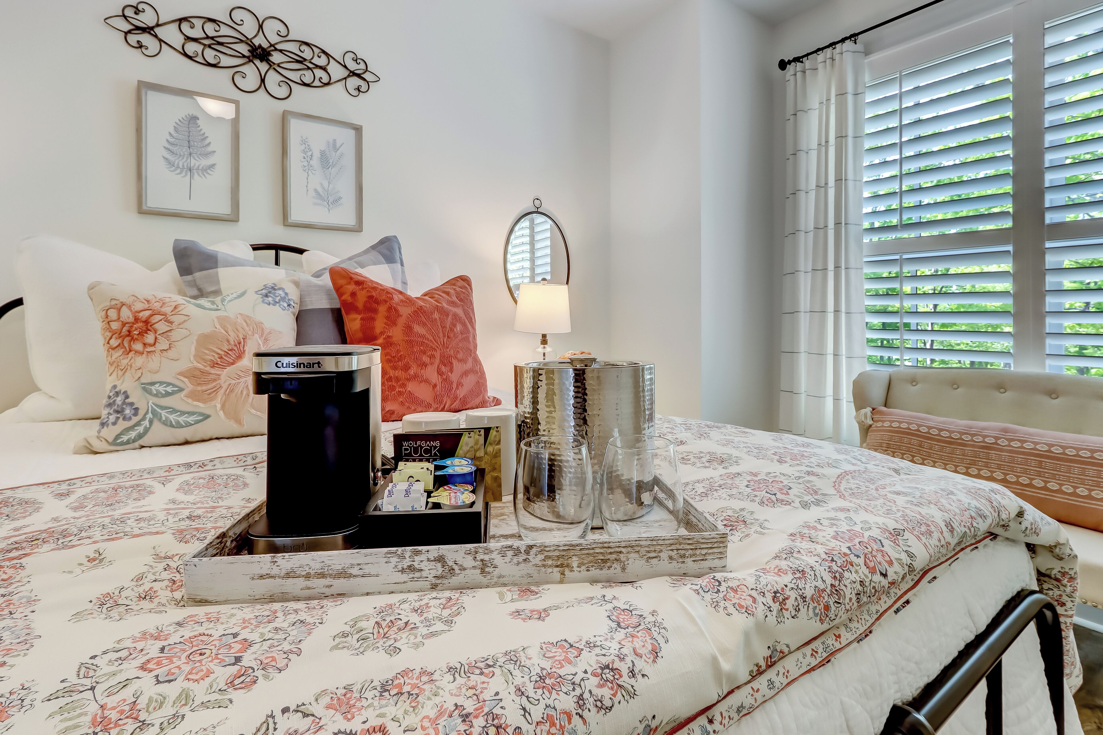 A neatly made bed with a floral bedspread, a bedside table with a lamp, and a window with blinds.