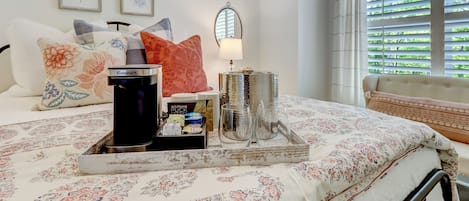 A neatly made bed with a floral bedspread, a bedside table with a lamp, and a window with blinds.