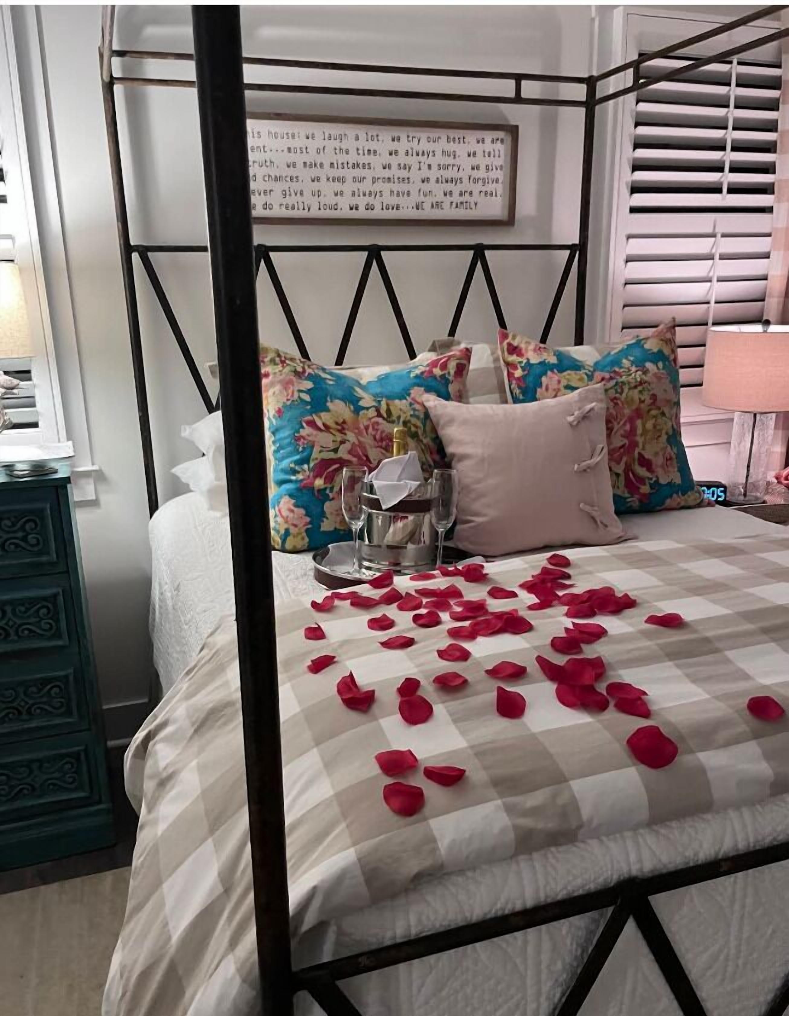 A four-poster bed with floral pillows, a bedside table with a lamp, and rose petals scattered on the bed.