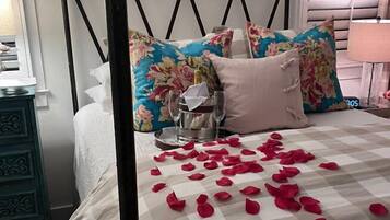 Uma cama de dossel com almofadas florais, uma mesinha de cabeceira com abajur e pétalas de rosa espalhadas pela cama.