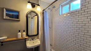 A bathroom with a white sink, mirror, and shower curtain. There is a framed picture on the wall and a window above the shower.